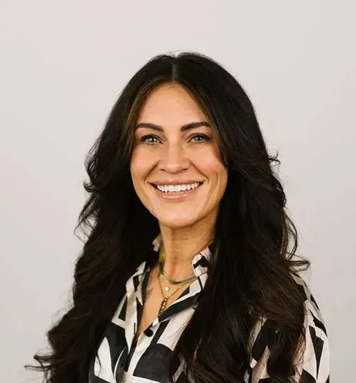 A smiling person with long, dark wavy hair wearing a patterned button-up shirt and layered necklaces against a grey wall.