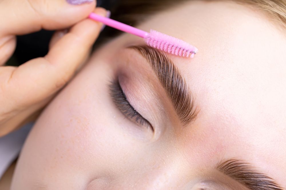 A hand uses a pink spoolie brush to groom and shape an eyebrow.