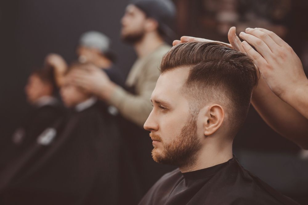A barber styles a man's hair in a salon while other clients wait in the background.