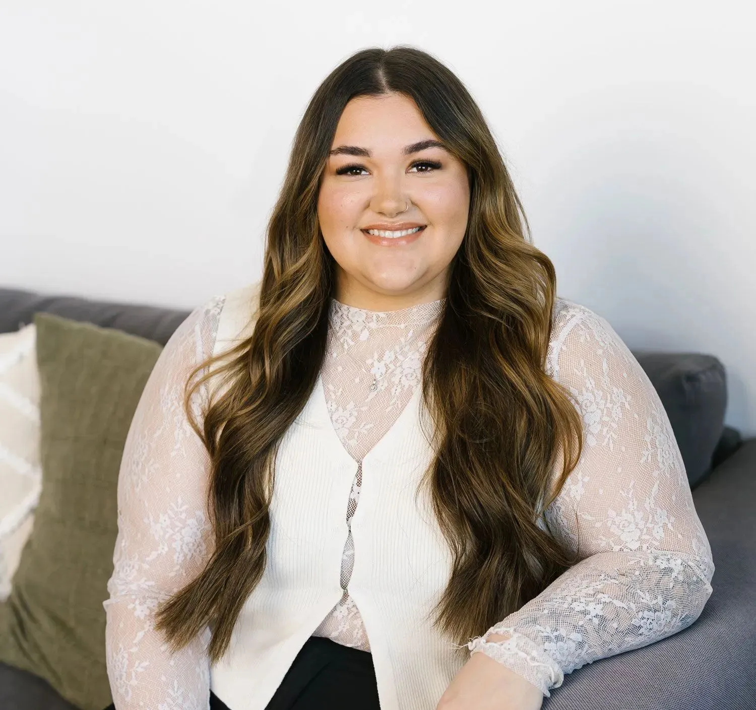 Smiling person with long, wavy brown hair, wearing a white lace-patterned top, sitting on a gray couch.