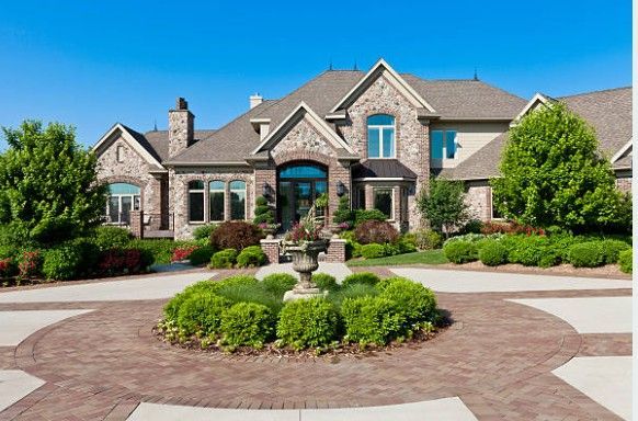Large stone house with circular driveway, manicured landscaping, and fountain.