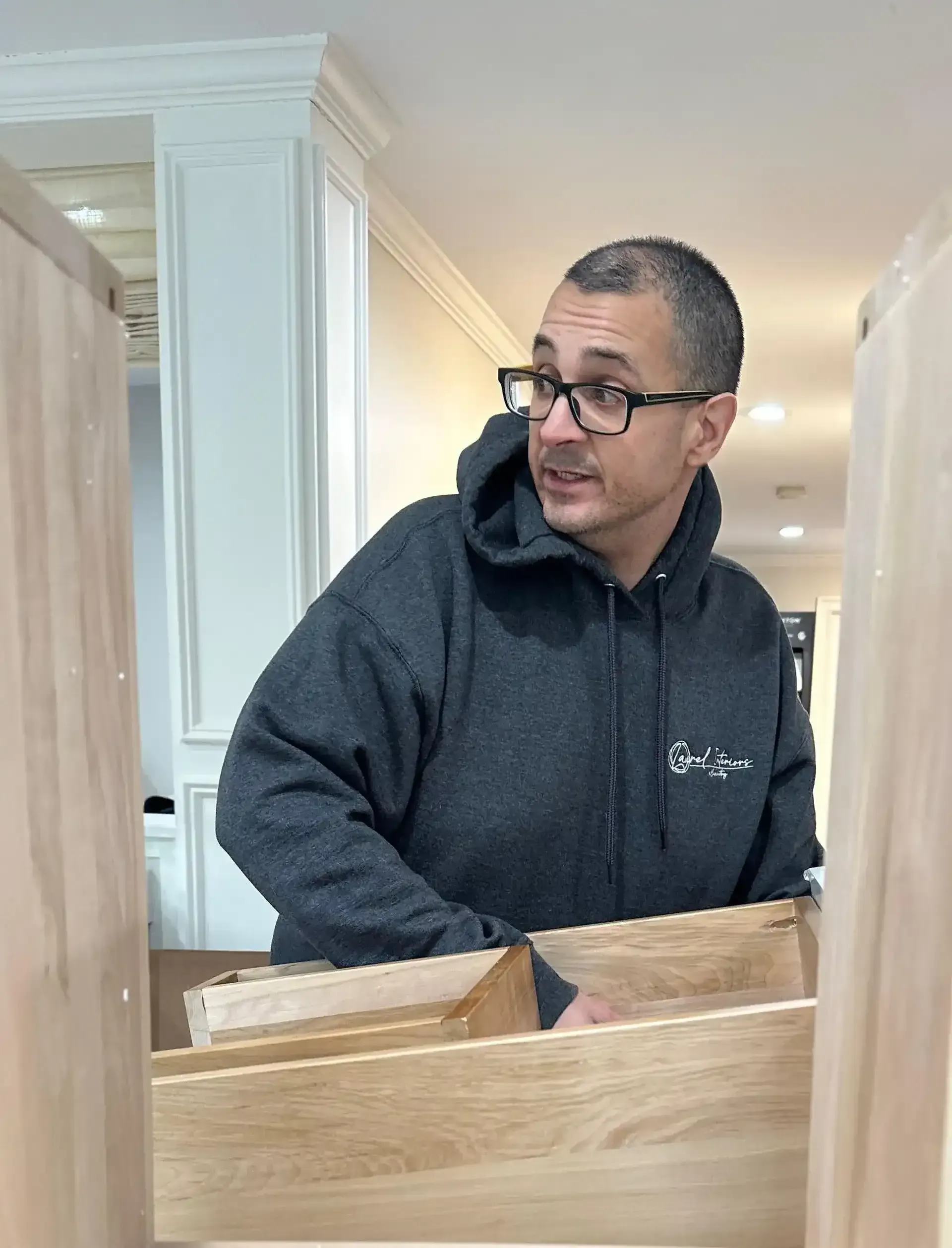 Person in a dark hoodie leaning over a wooden cabinet in a bright kitchen, looking to the side.