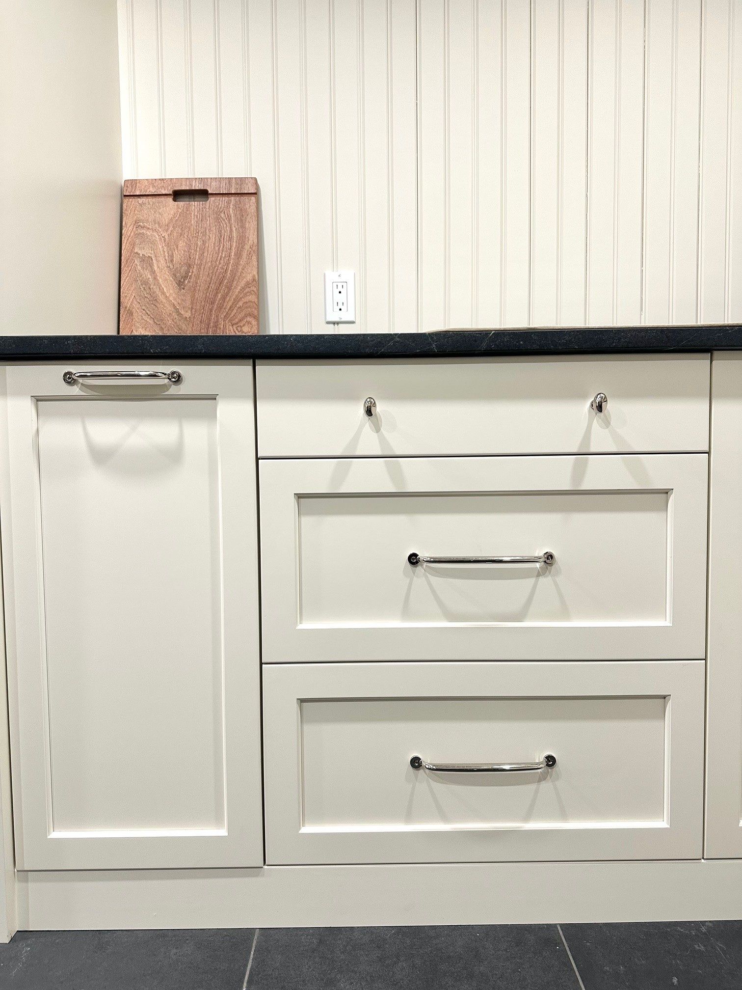 White kitchen cabinets with black countertop and wood cutting board against a paneled wall.