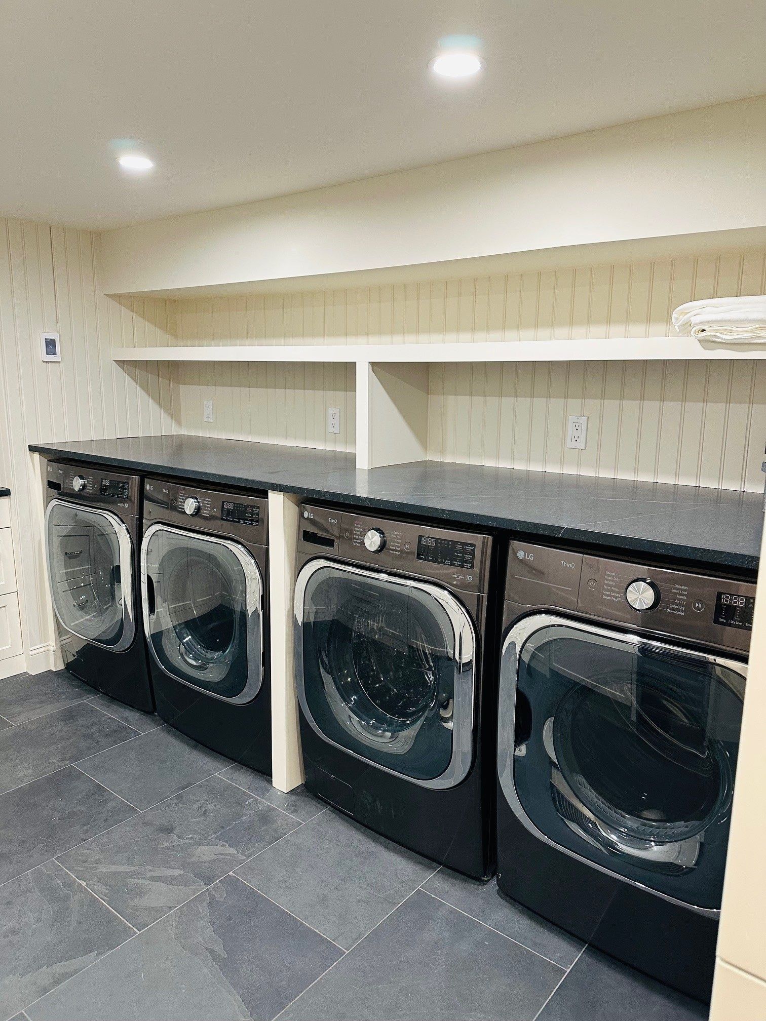 Row of front-loading washers in a beige laundry room with dark tile flooring and shelves above.