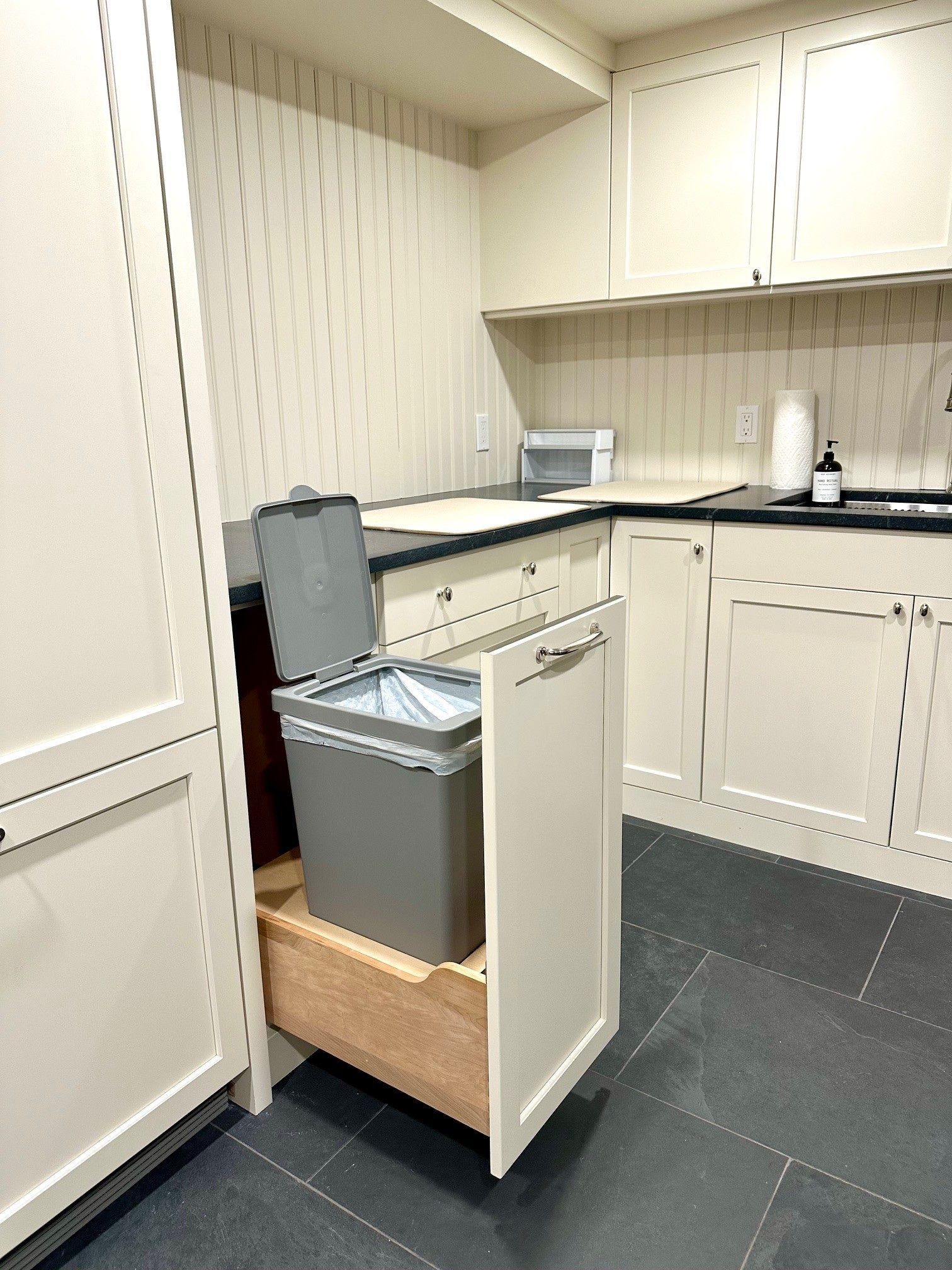 Kitchen with a pull-out trash bin drawer open beside white cabinets and black countertop.