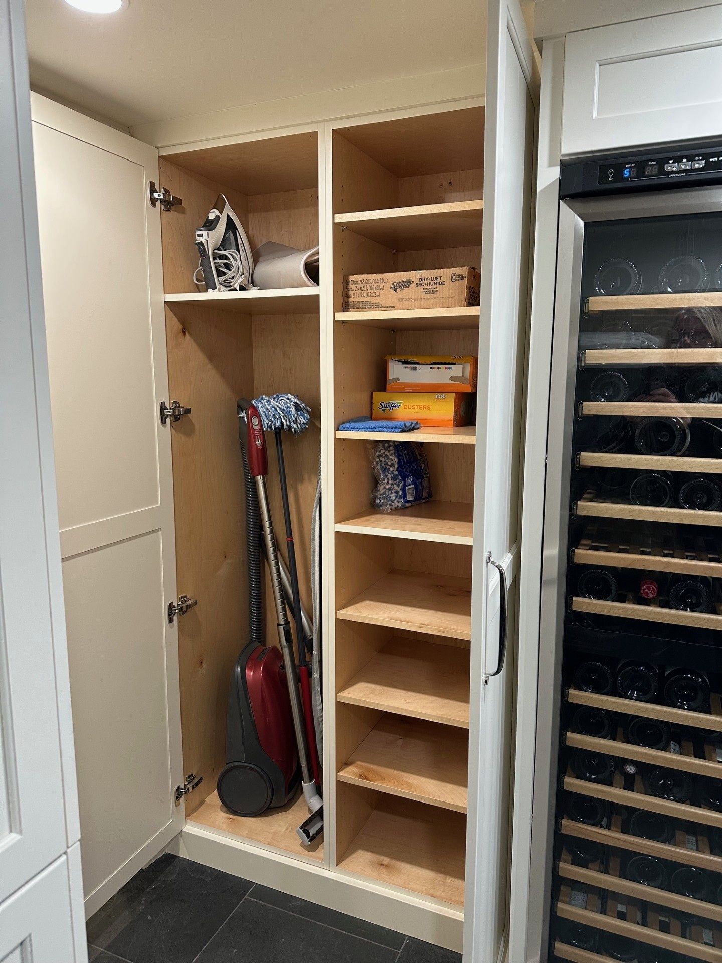 Open pantry closet with shelves, broom, dustpan, and cleaning supplies beside a wine fridge