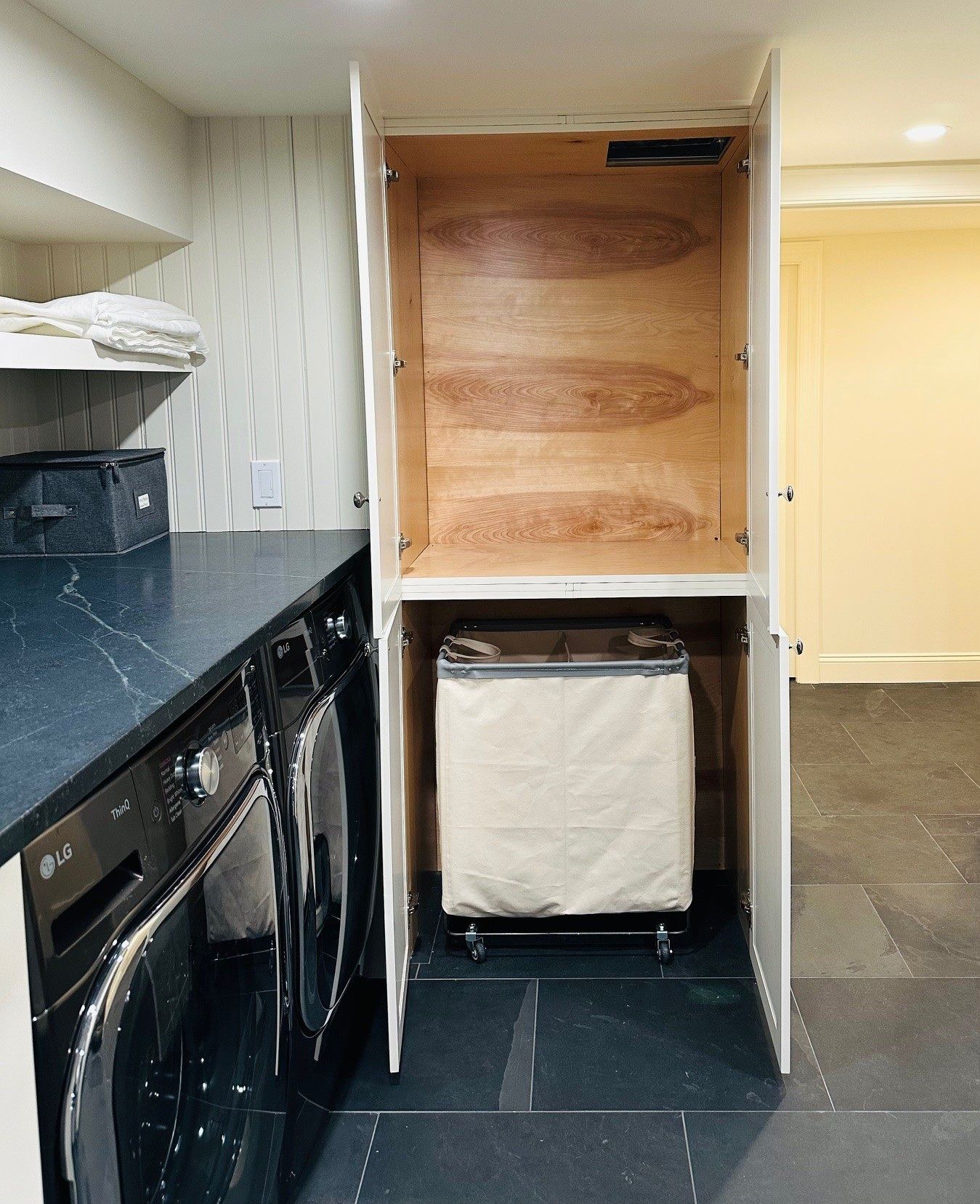 Narrow kitchen alcove with stacked washer and dryer under open wooden cabinets and black countertops.