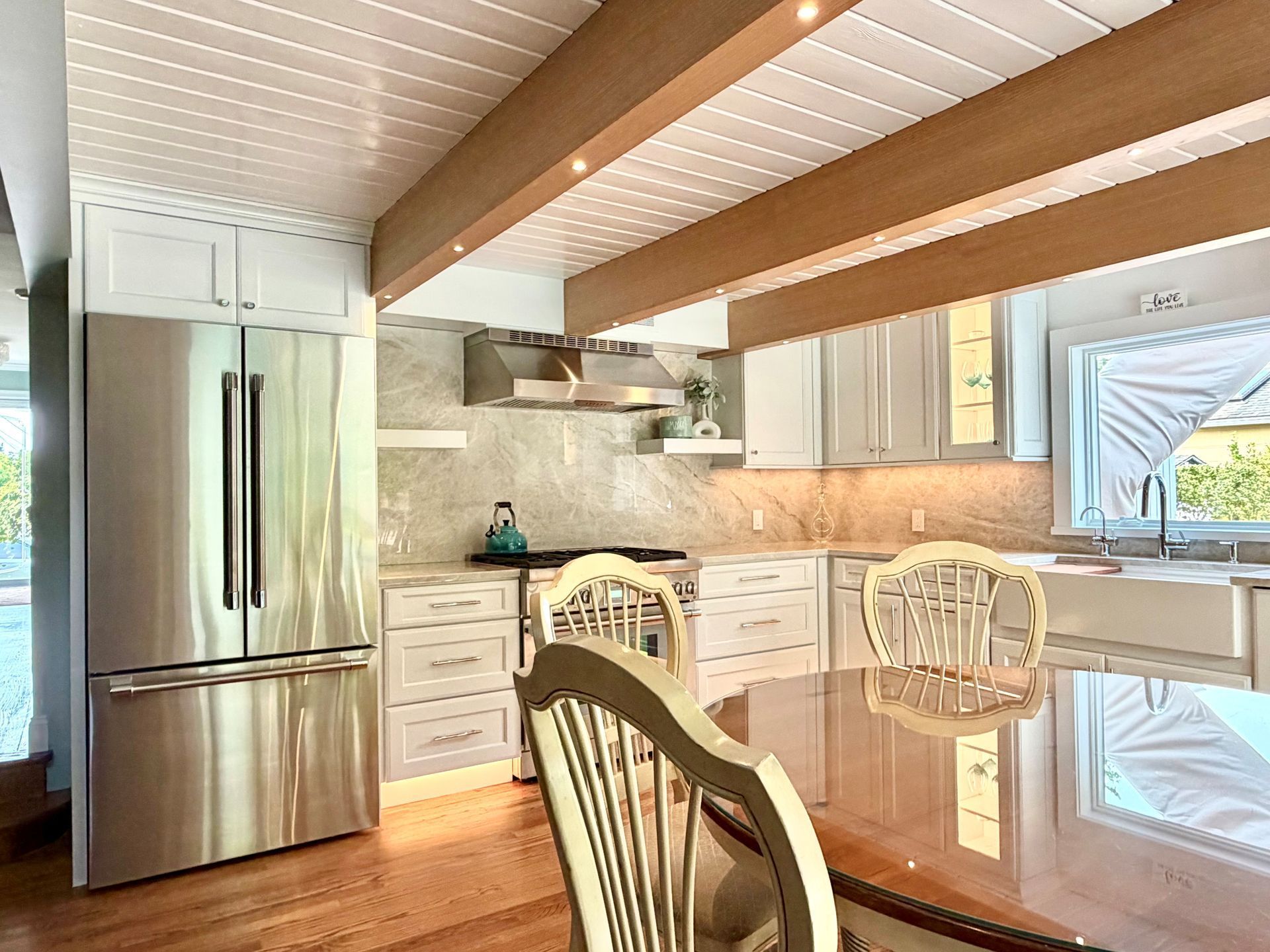 Bright modern kitchen with stainless steel fridge, white cabinets, wood beams, and glass dining table
