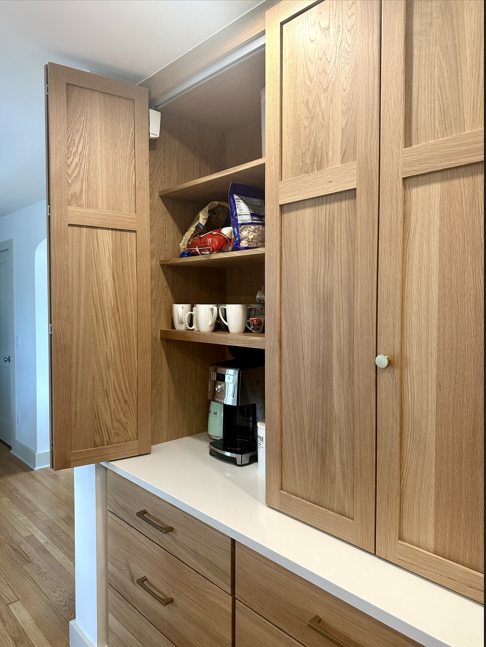 Open kitchen cabinet with wooden doors, shelves holding mugs, snacks, and a coffee maker over a countertop.
