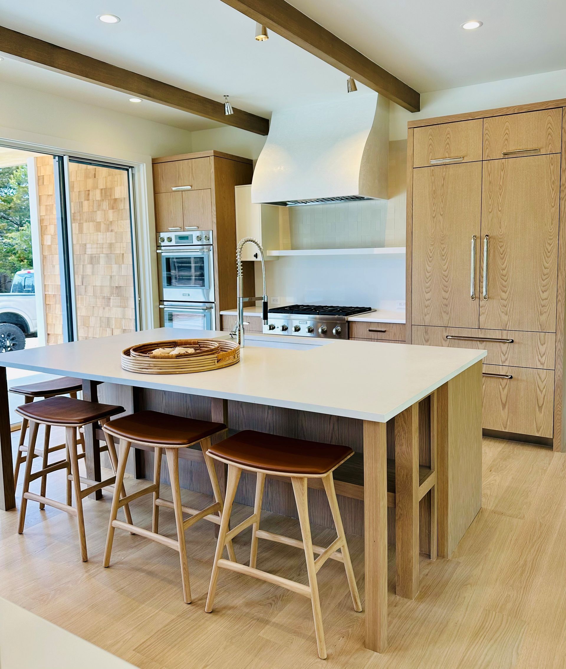 Modern kitchen with a large white island, wooden stools, and light wood cabinetry with built-in appliances