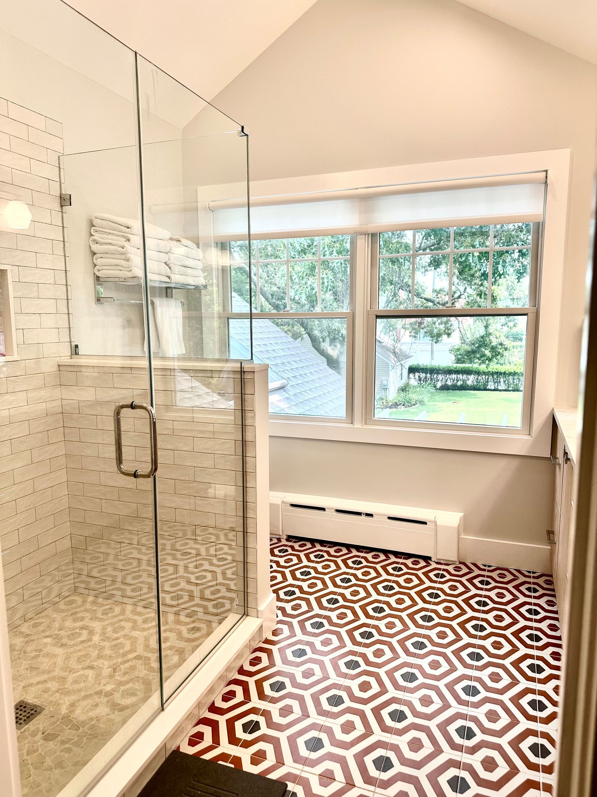Glass-enclosed shower beside a window in a bright bathroom with patterned tile floor.