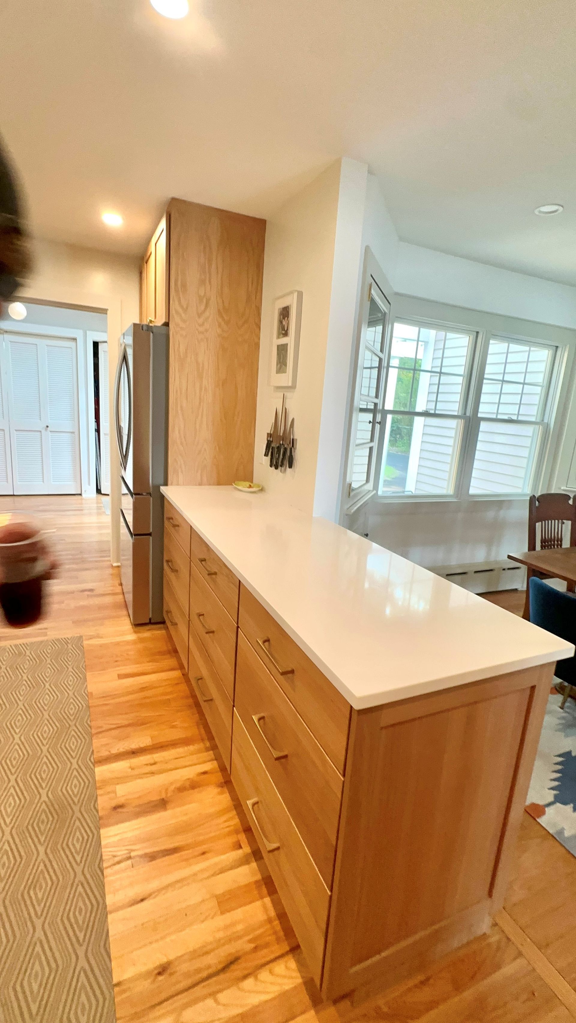 Bright kitchen with long white island, wood cabinets, hardwood floors, and windows at the end.