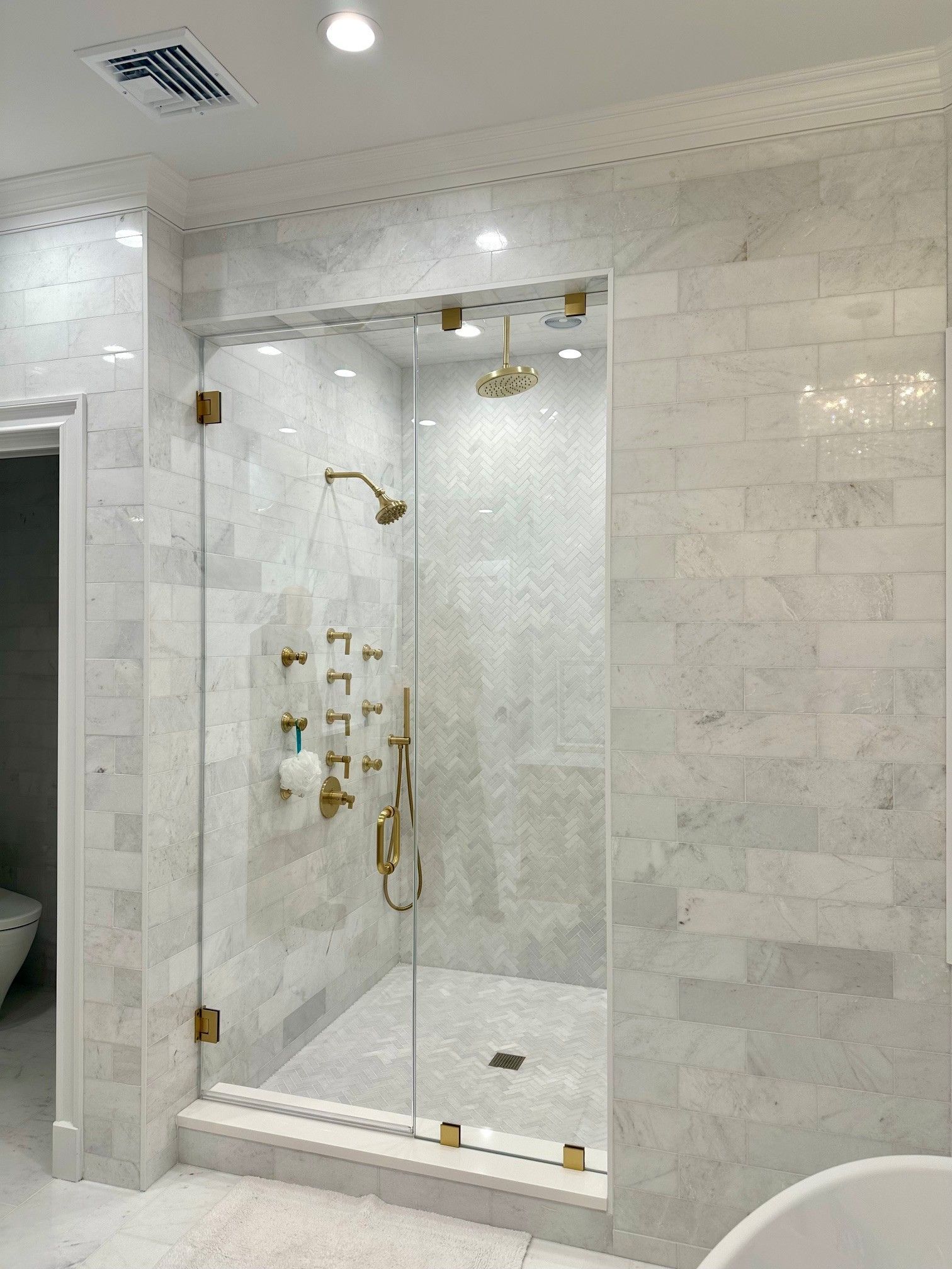 Modern white marble shower with glass door, brass fixtures, and a built-in bench in a tiled bathroom