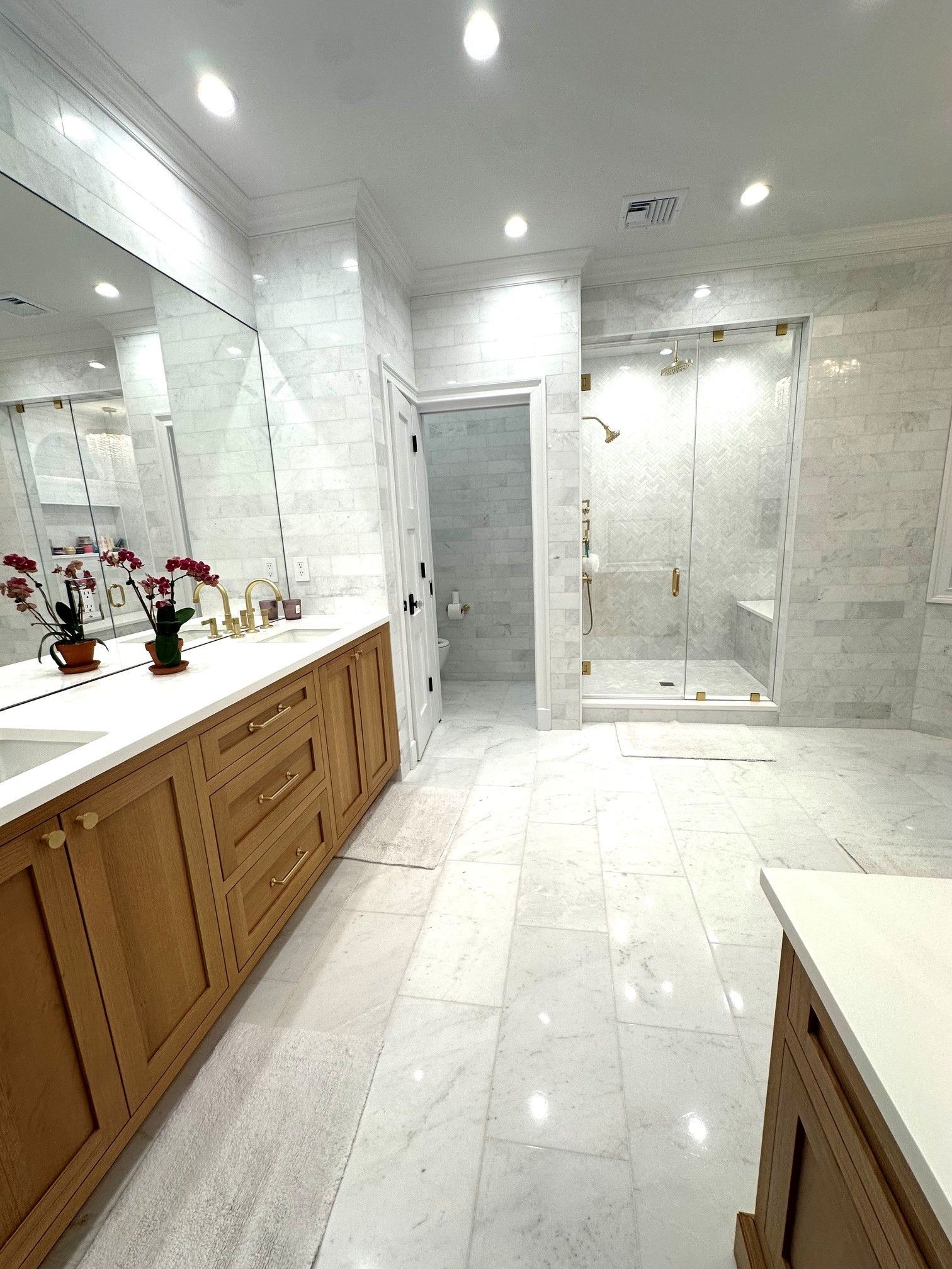 Bright white marble bathroom with wood vanity, large mirror, glass shower, and recessed ceiling lights