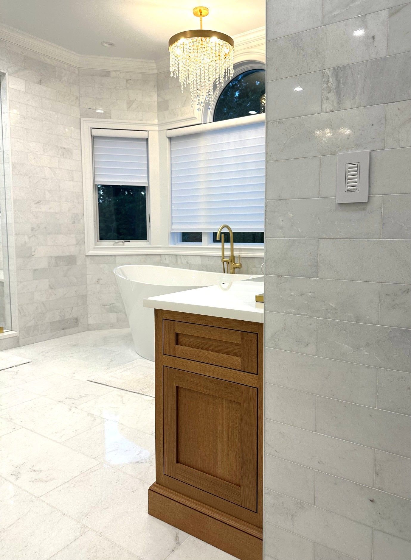Bright marble bathroom with a wooden vanity, gold faucet, chandelier, and window above a freestanding tub.
