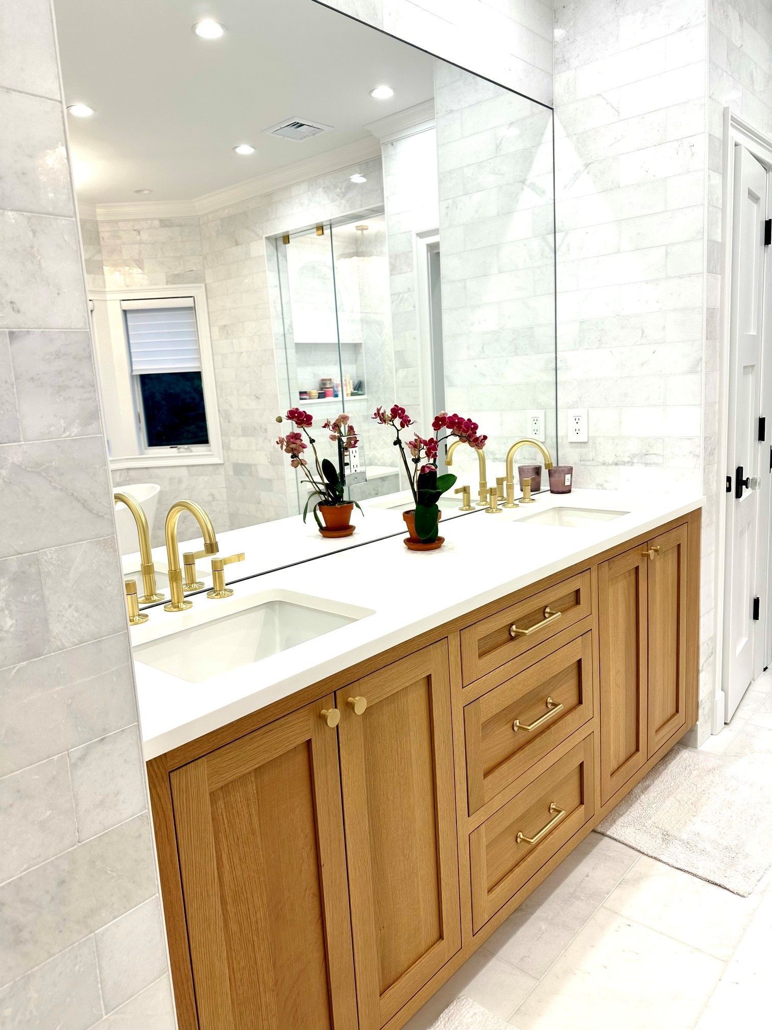 Bright bathroom with marble walls, wood double vanity, gold faucets, and flowers on the countertop.
