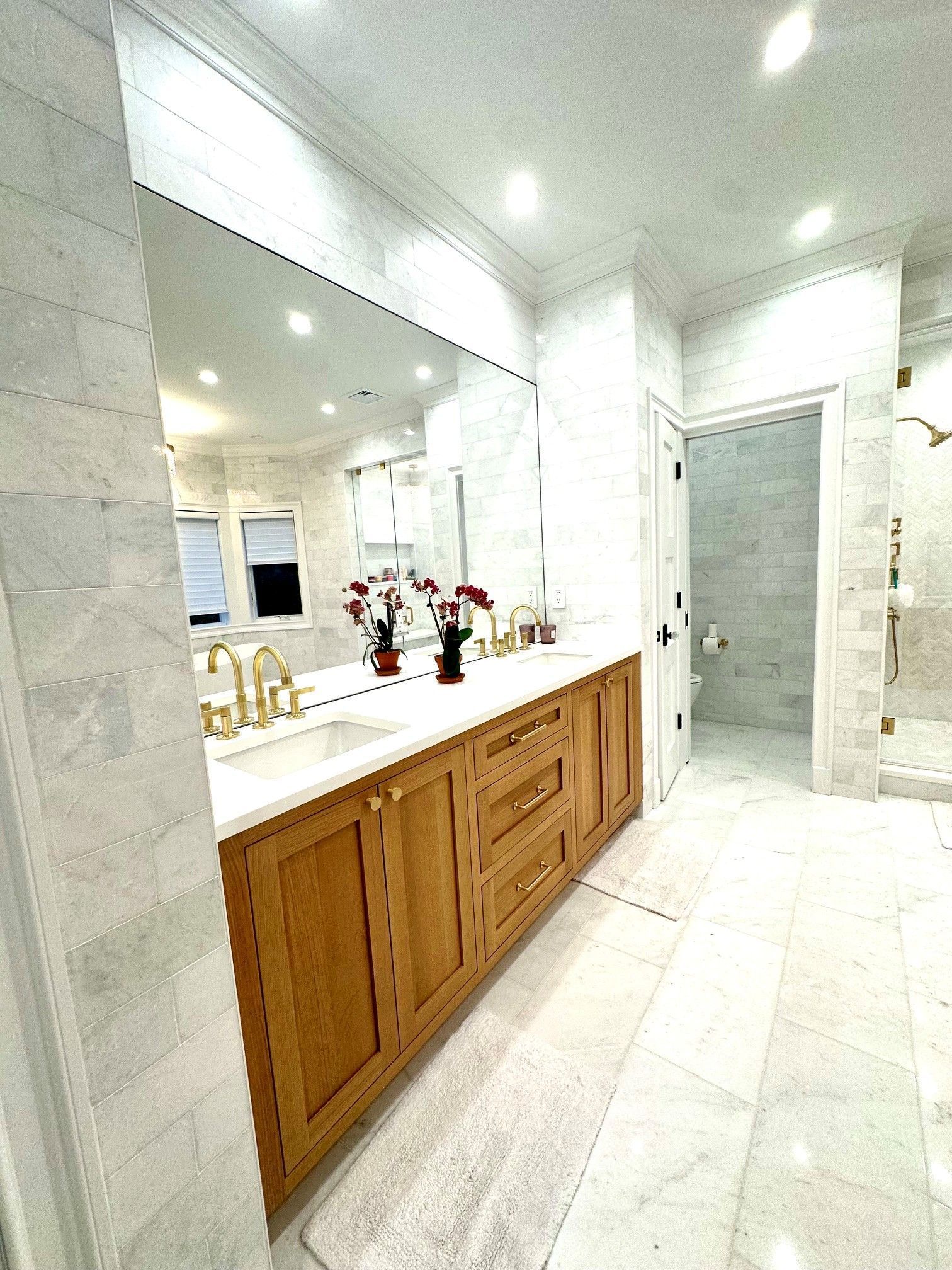 Bright white bathroom with double wood vanity, large mirror, marble tile, and glass shower door.
