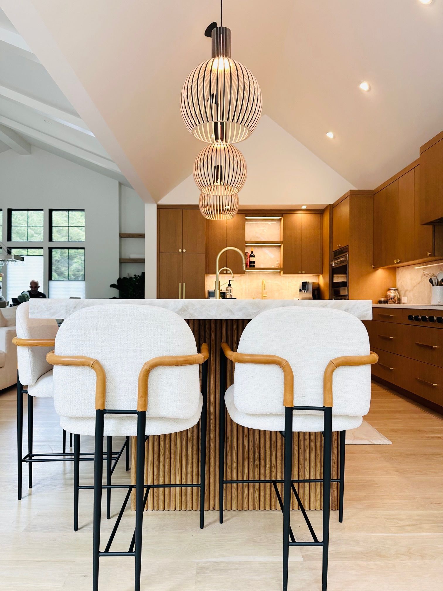 Modern kitchen with wood cabinetry, marble island, and two white upholstered bar stools under pendant lights