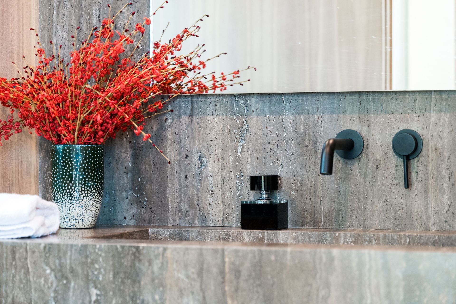 Modern bathroom sink with stone backsplash, black fixtures, and a vase of bright red flowers