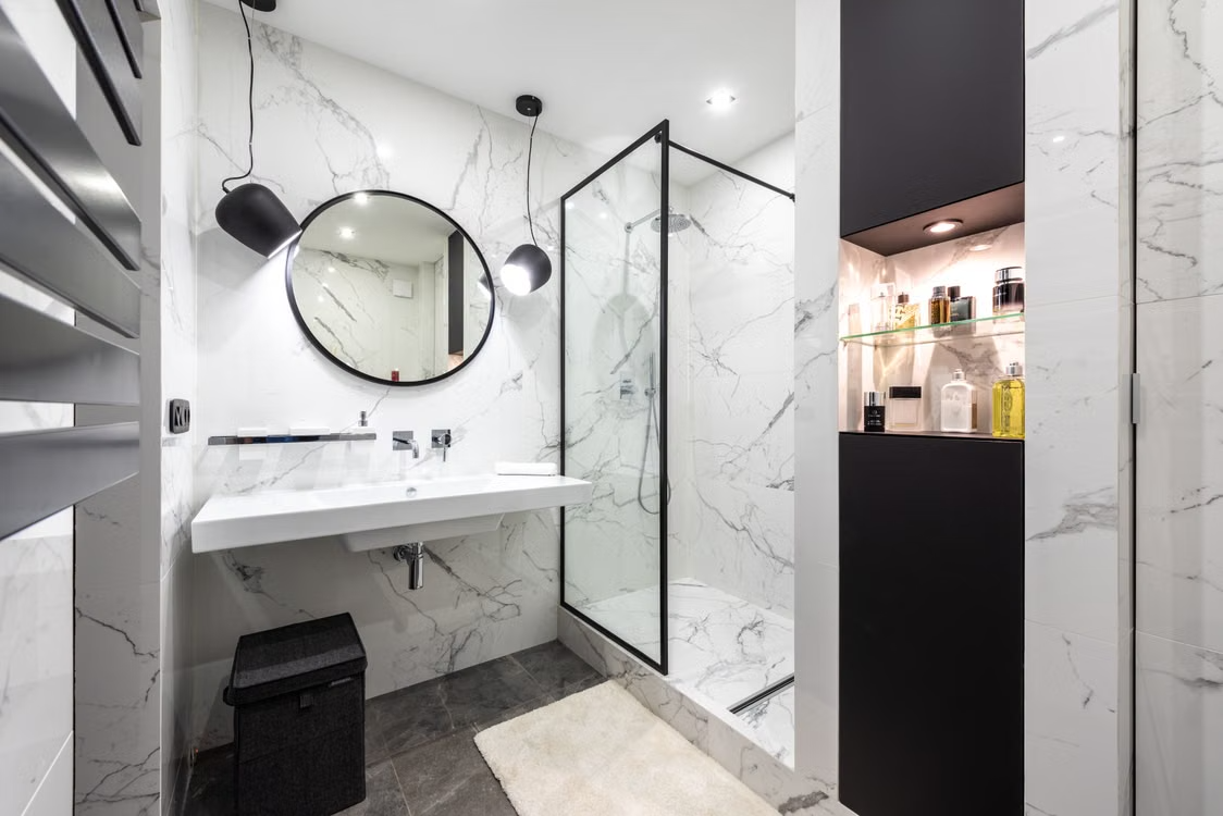 Modern white marble-tiled bathroom with round mirror, vanity sink, glass shower, and black accents