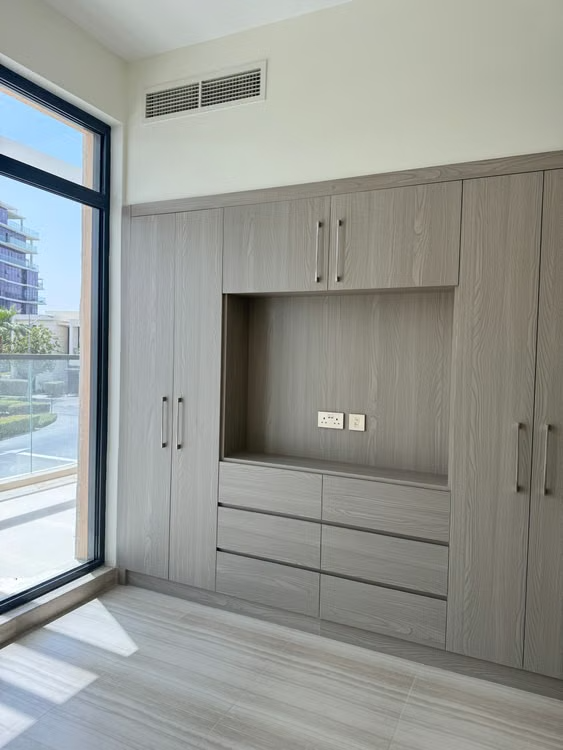 Built-in gray wardrobe with drawers beside a large window in a bright empty room