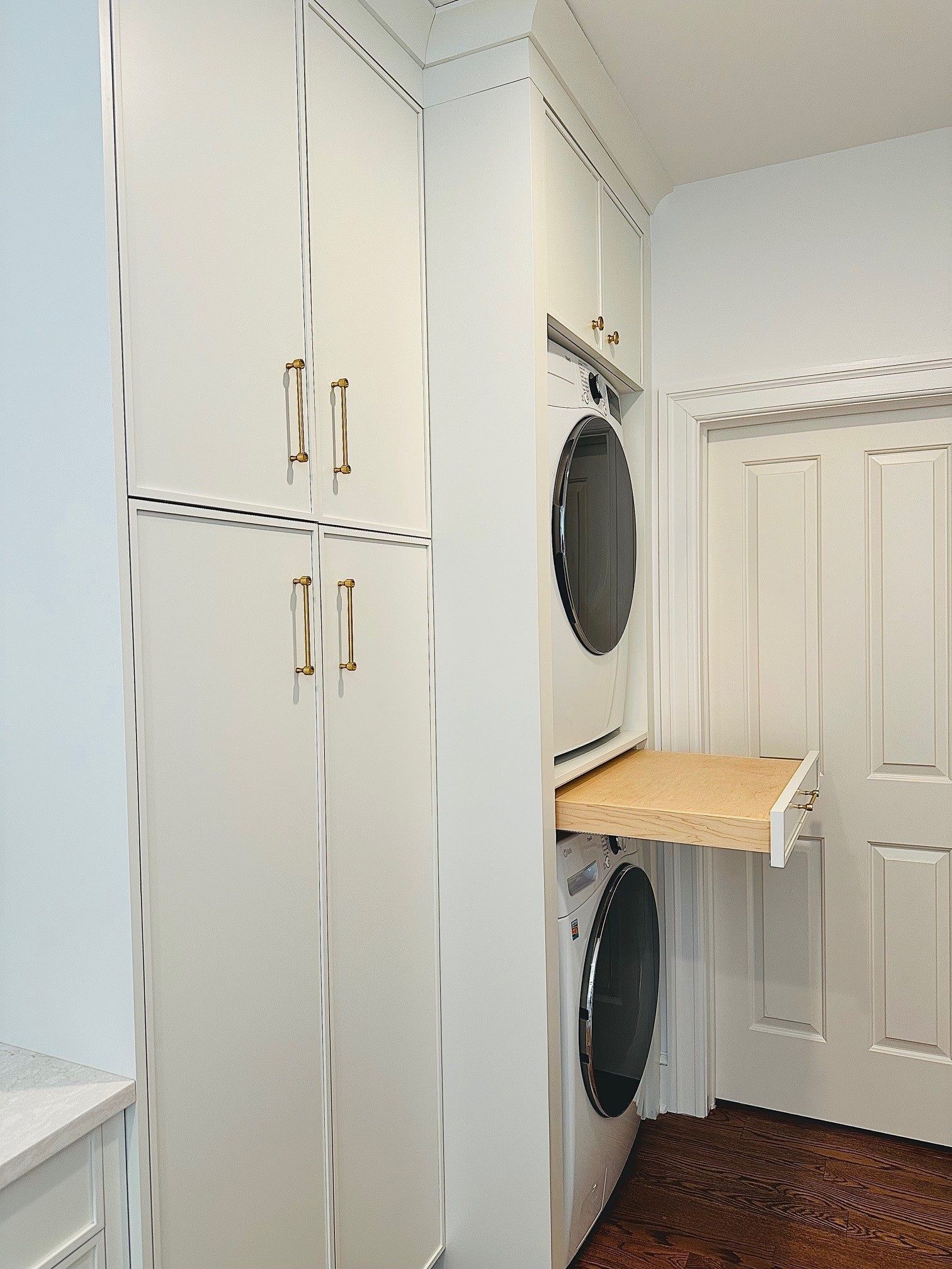 Stacked washer and dryer beside white cabinets in a narrow laundry room with a small folding shelf