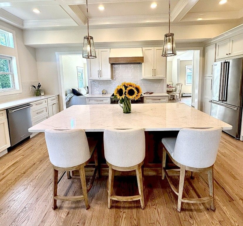 Bright modern kitchen with large island, three white stools, pendant lights, and a vase of sunflowers.