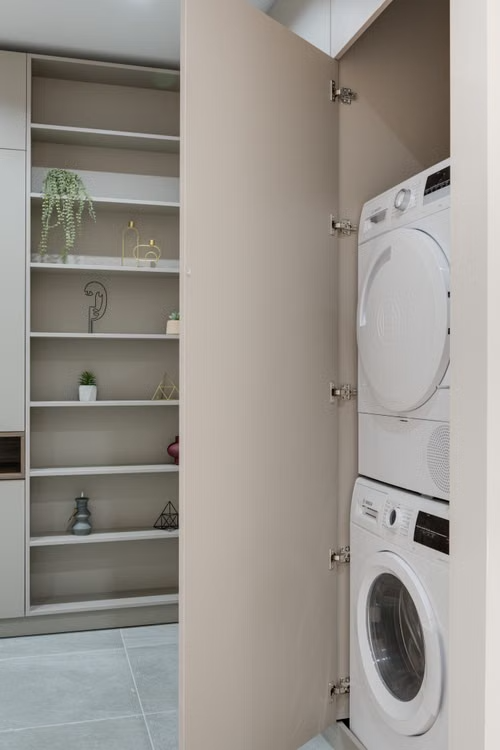 Laundry closet with stacked washer and dryer beside empty shelves in a beige room