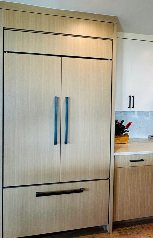 Tall light-wood kitchen cabinet with blue handles beside a white countertop and cabinets.