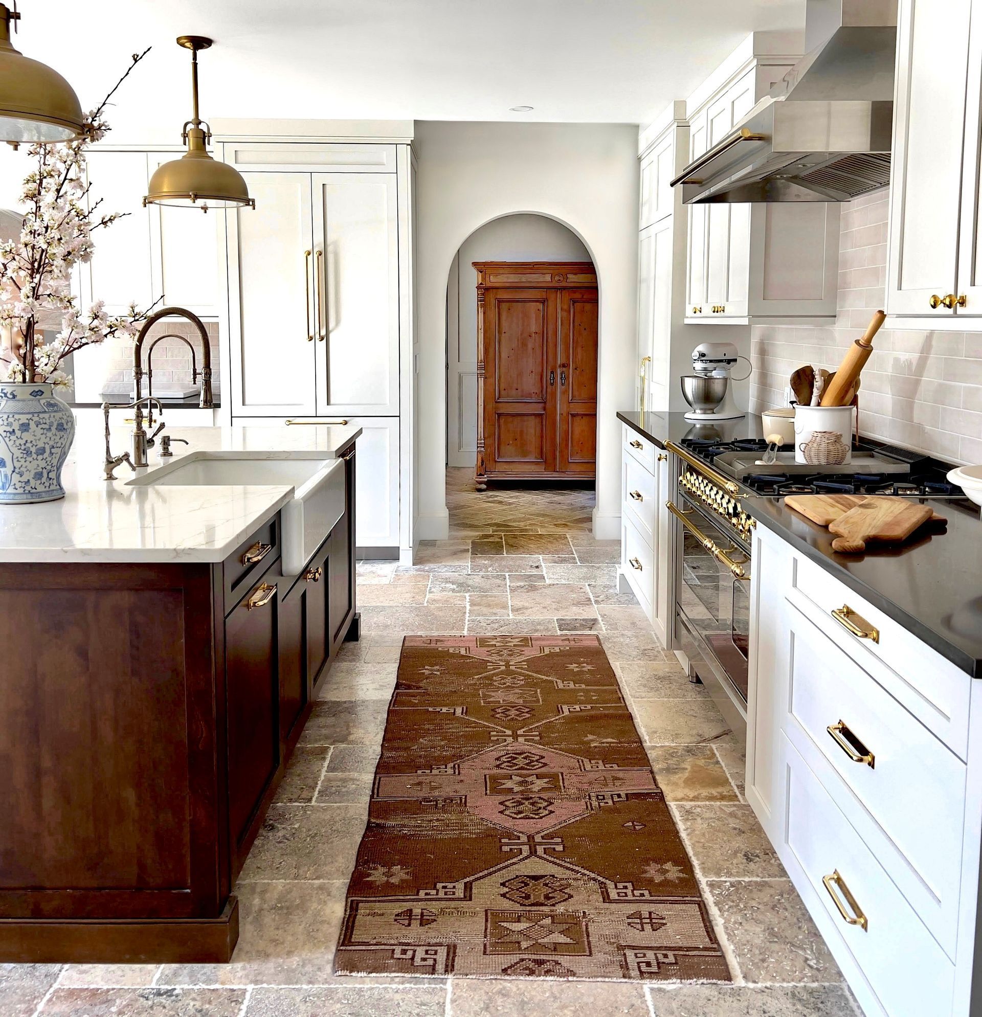 Bright white kitchen with wood island, brass fixtures, patterned runner, and rustic wooden door at the end.