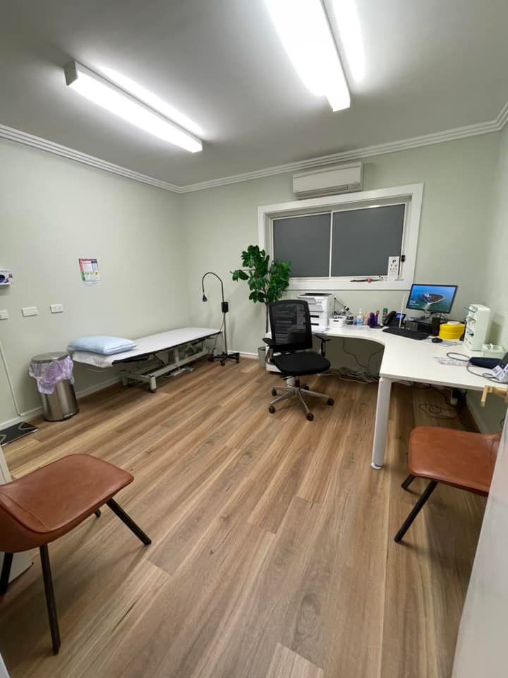 Small medical exam room with exam table, desk, office chair, and overhead fluorescent lights