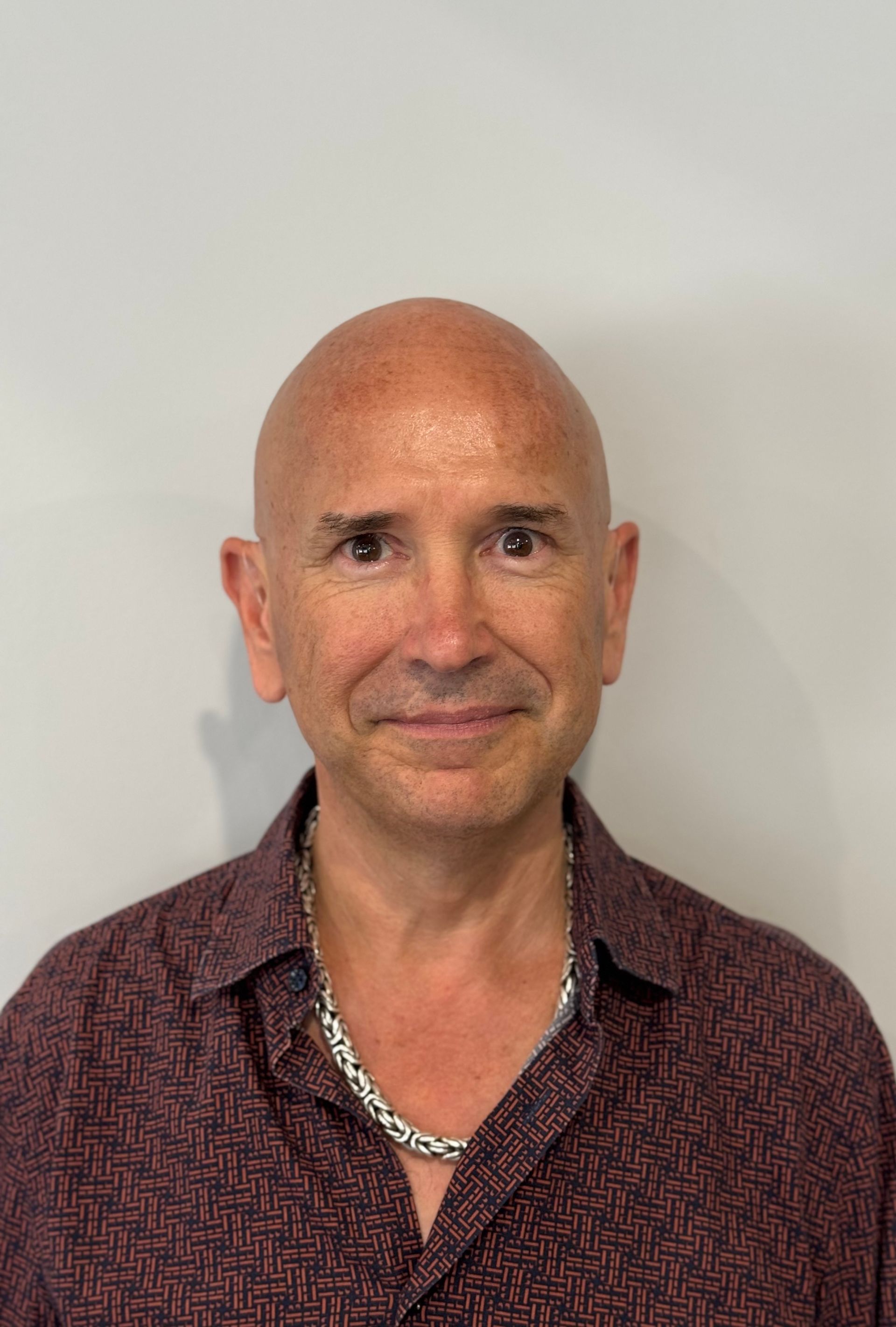 Smiling bald man in a patterned maroon shirt and silver chain against a plain white wall