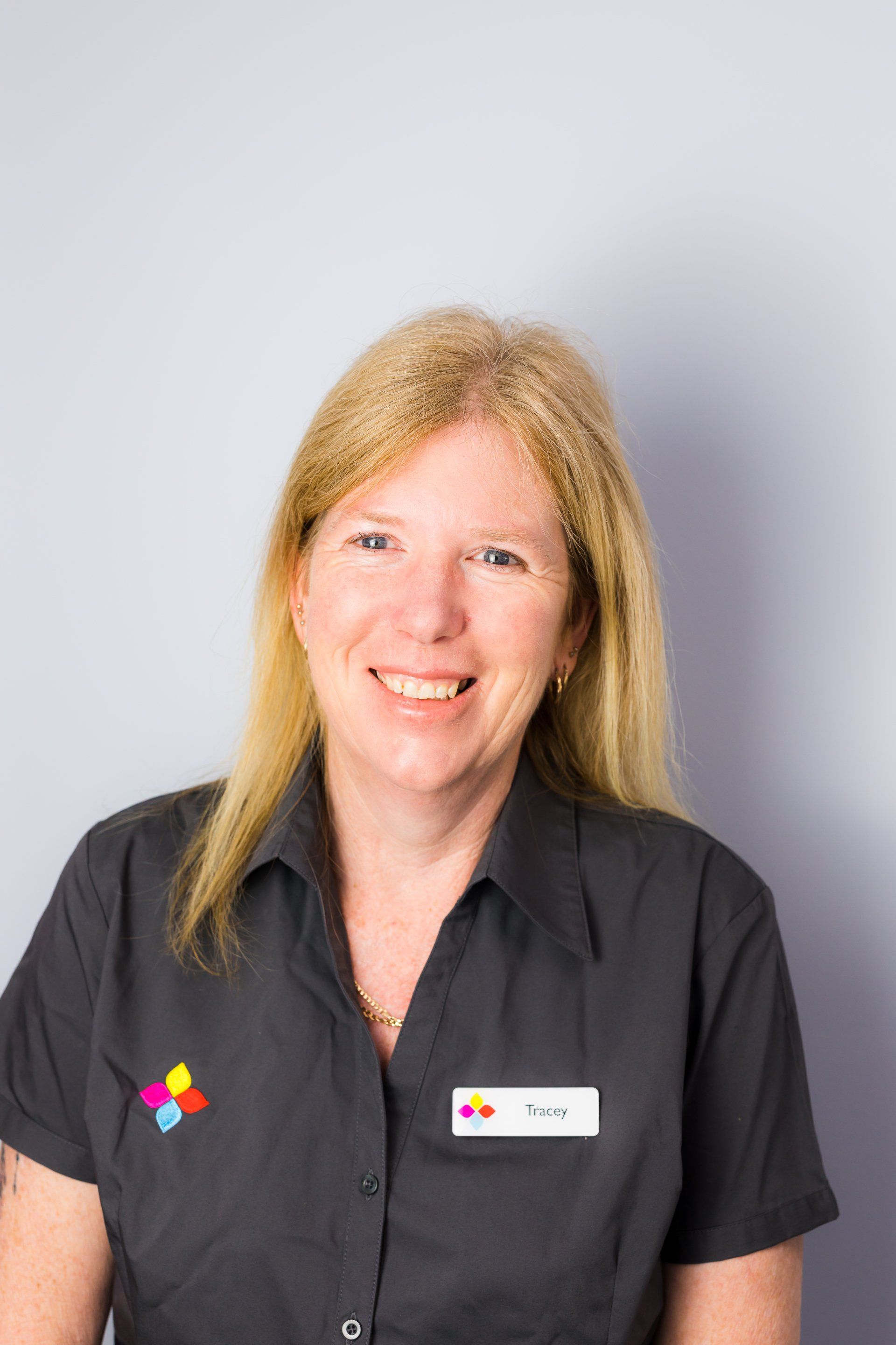 Smiling woman in a black shirt with a logo and name tag against a light gray background