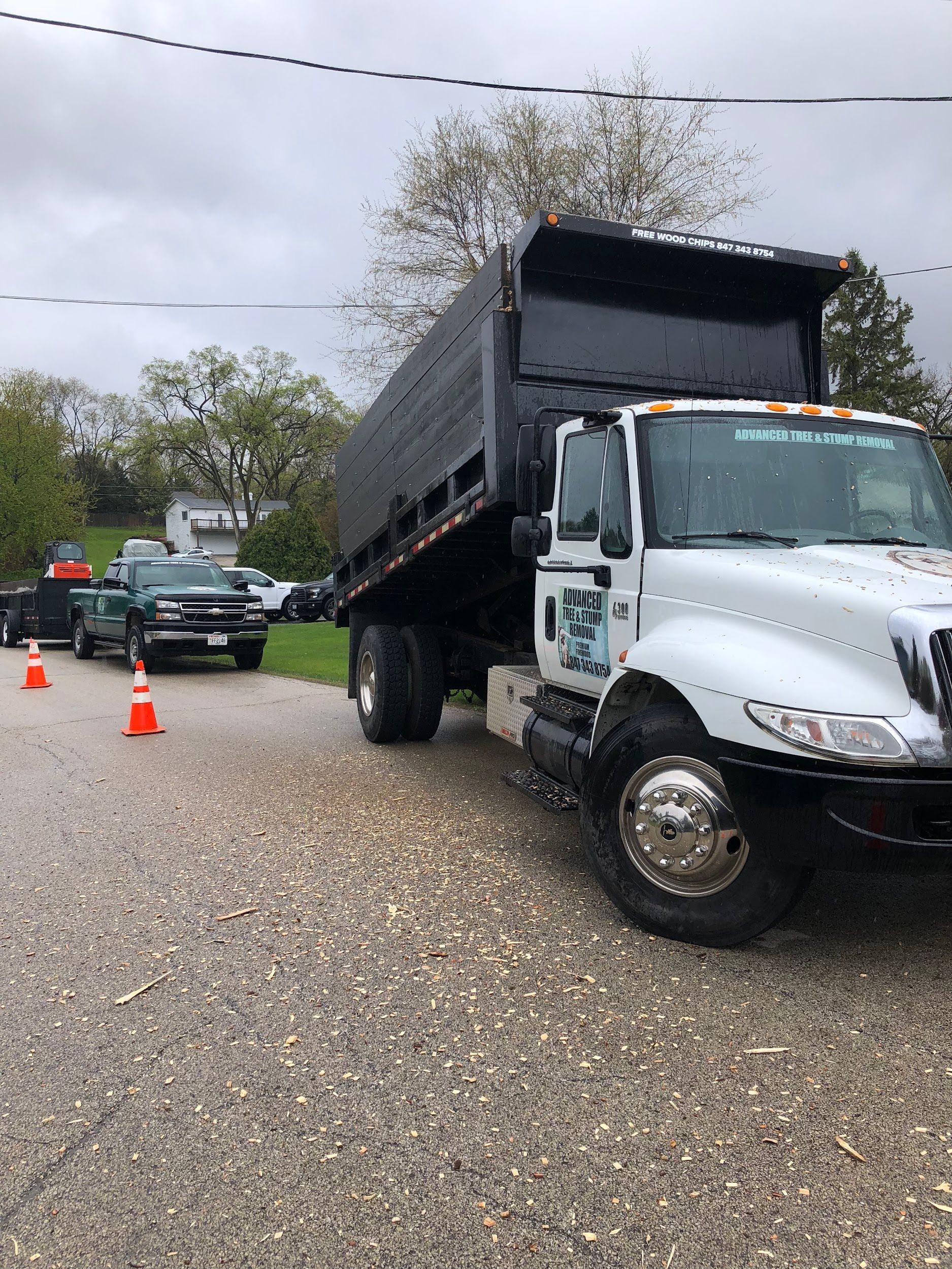 Dump truck with raised bed, parked on gravel road with orange cones. Green truck and houses in background.