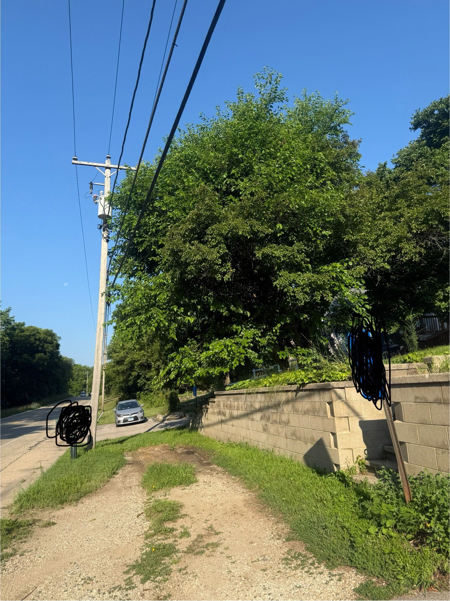 Gravel road next to a concrete wall and lush green trees under a blue sky, power lines on the left.