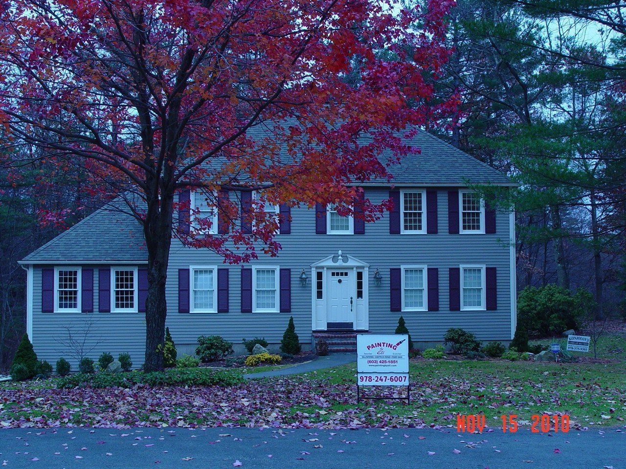 Two-story gray house with white trim, red autumn tree, and a yard sign in front at dusk