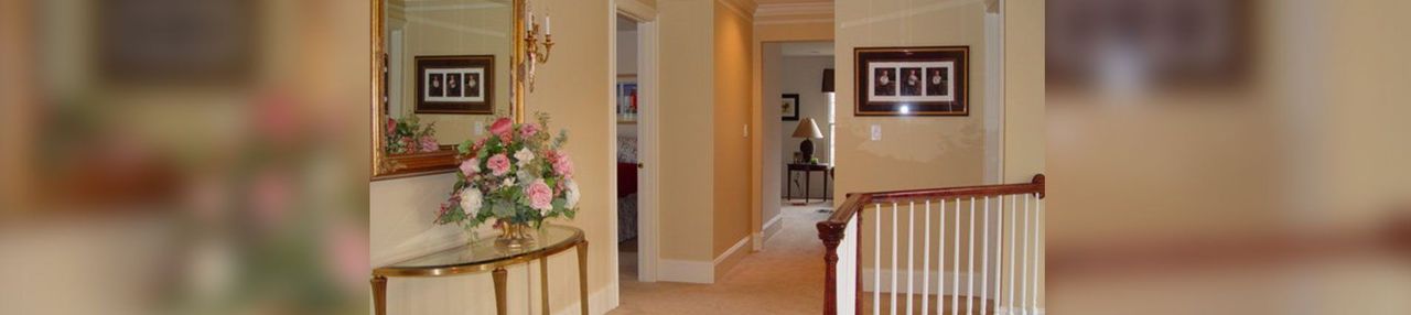 Warm hallway with framed photos, a floral arrangement, and a wooden staircase railing in a home interior Warm hallway with framed photos, a floral arrangement, and a wooden staircase railing in a home interior
