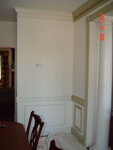 White paneled wall beside a doorway, with a chair and a partially visible room interior. White paneled wall beside a doorway, with a chair and a partially visible room interior.
