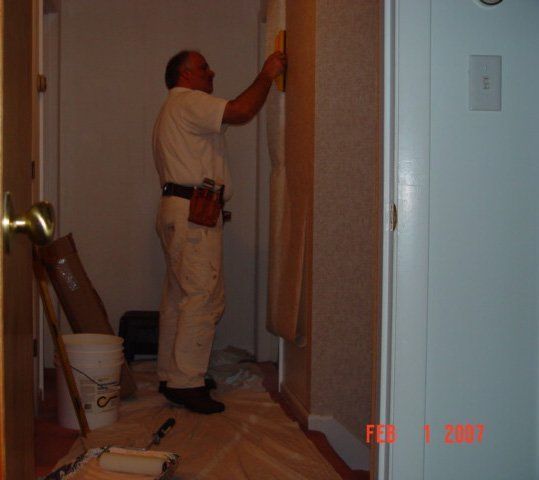 Worker standing on a floor in a hallway, painting or trimming near a doorway with tools on the floor.