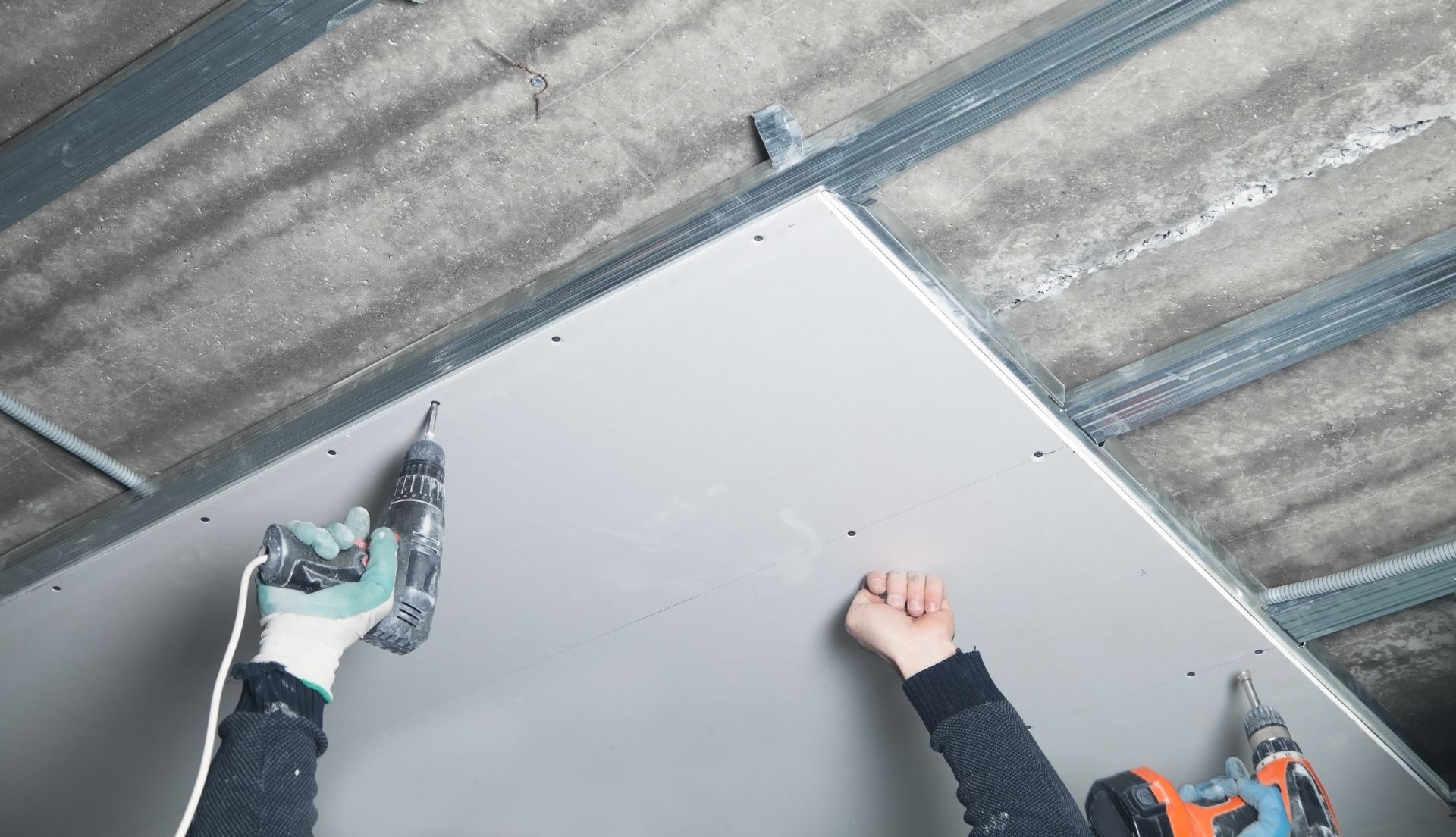 Two workers installing a gray ceiling panel with power drills under a metal roof