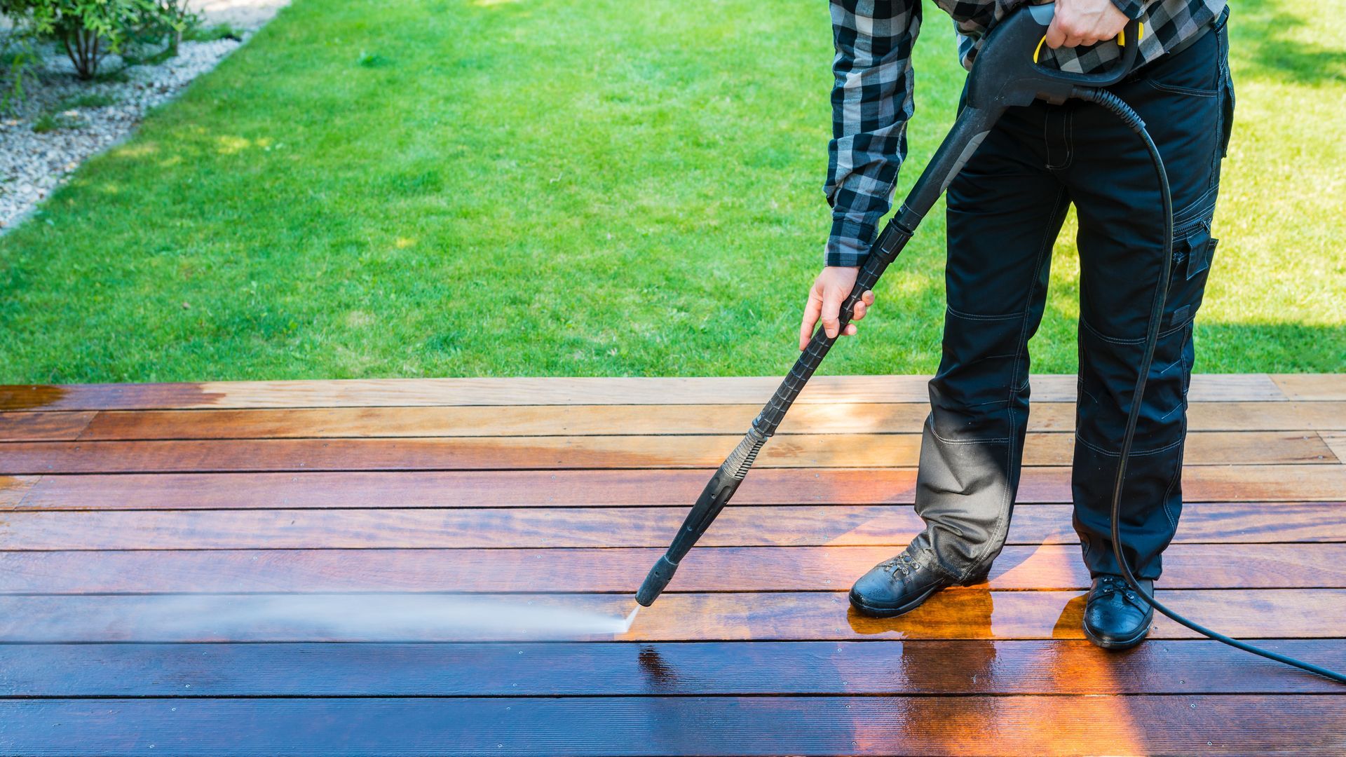 Person power-washing a wet wooden deck beside a green lawn Person power-washing a wet wooden deck beside a green lawn