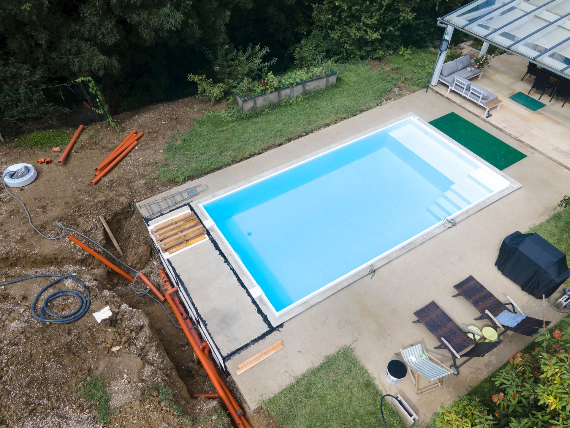 Aerial view of a finished in-ground pool and patio with leftover construction pipes in the yard