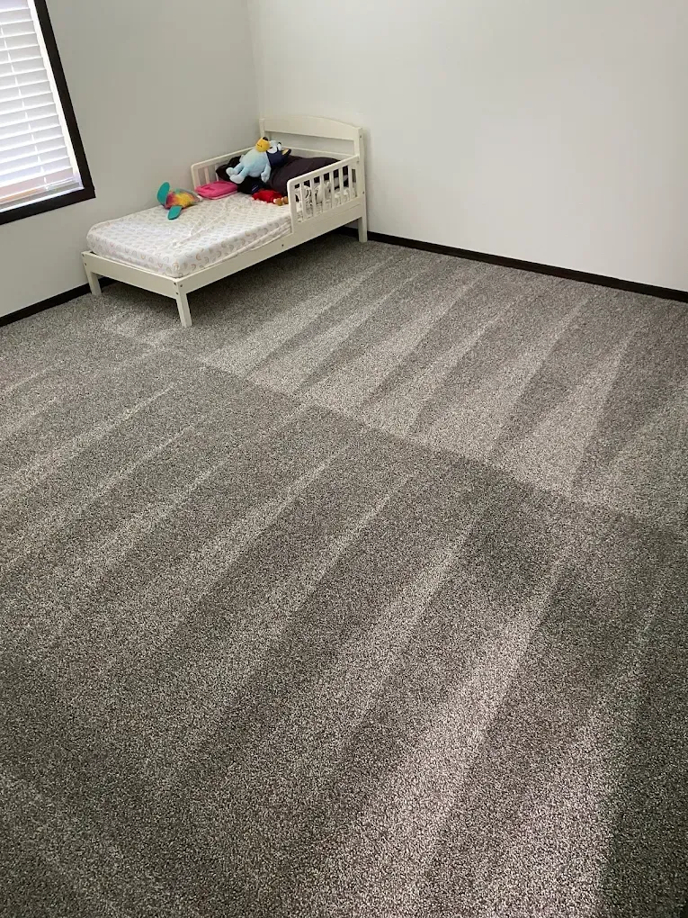 A small child's bed with stuffed animals sits in the corner of a room with gray, carpeted floors and a window.