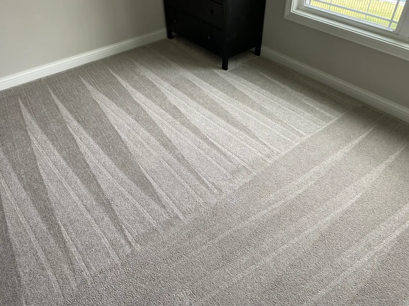 Gray carpet with a pattern of triangular vacuum tracks in a room with a black piece of furniture and a window.