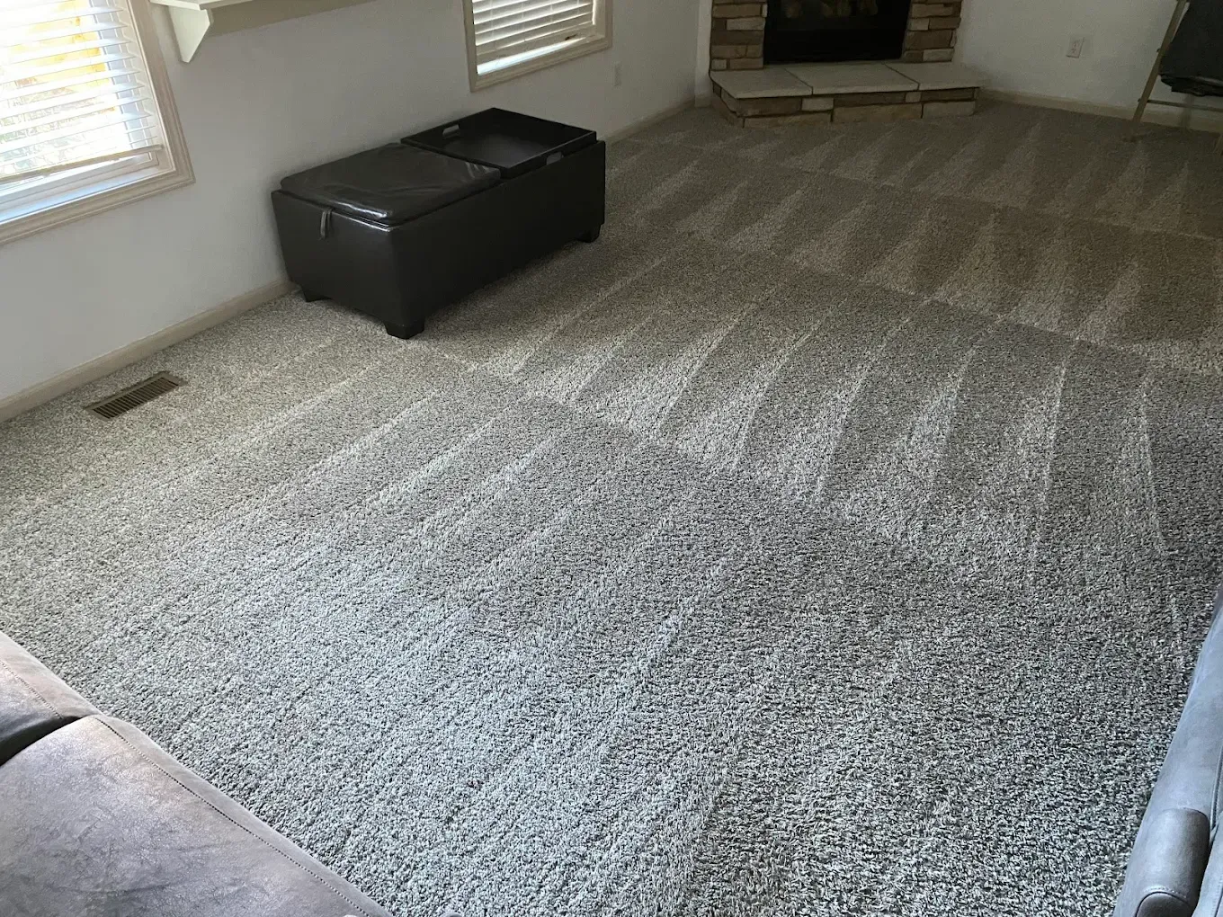 A living room with light gray patterned carpet, a dark leather storage ottoman, and a corner stone fireplace.