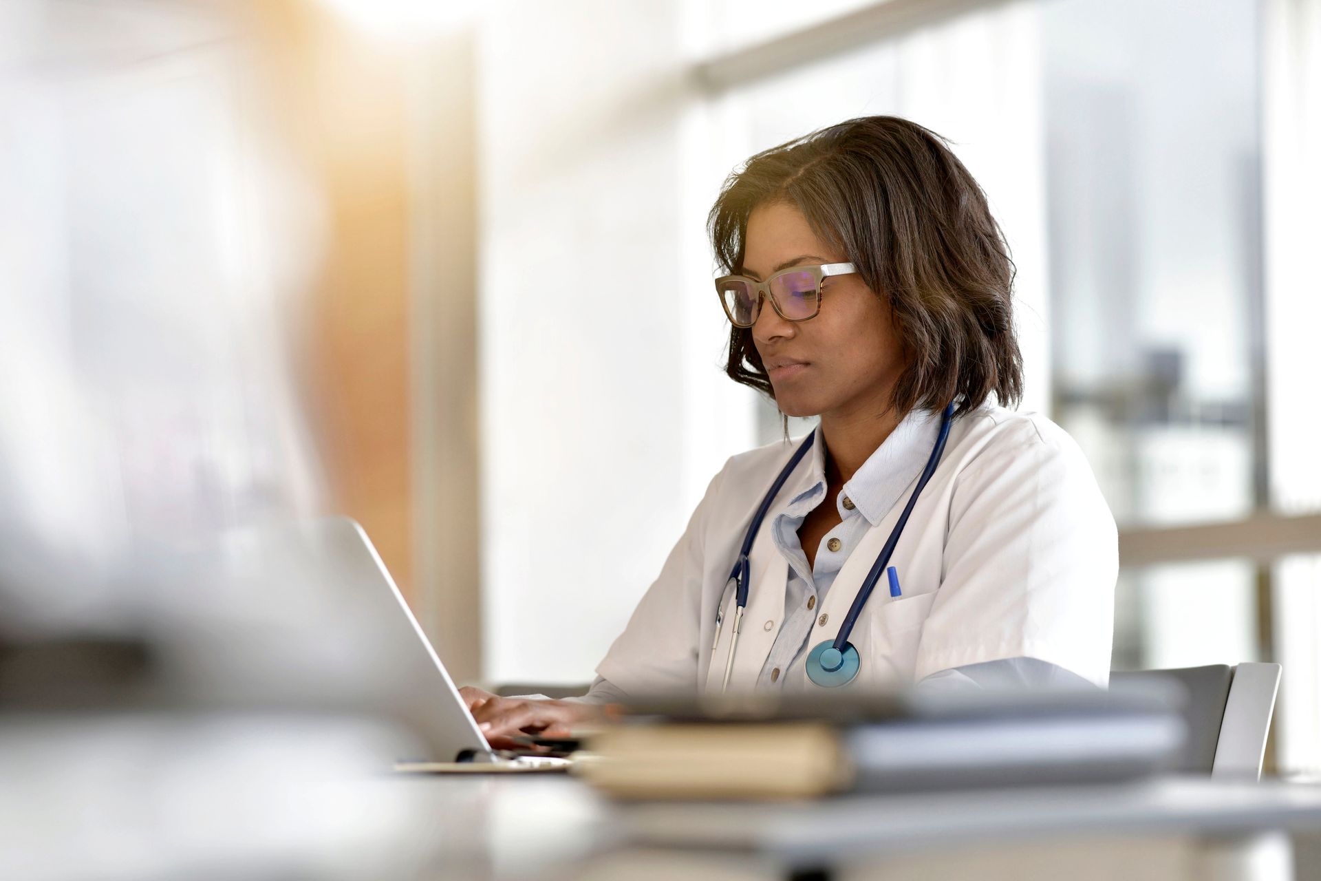 Médecin noir portant des lunettes et un stéthoscope tapant sur un ordinateur portable dans un bureau lumineux.
