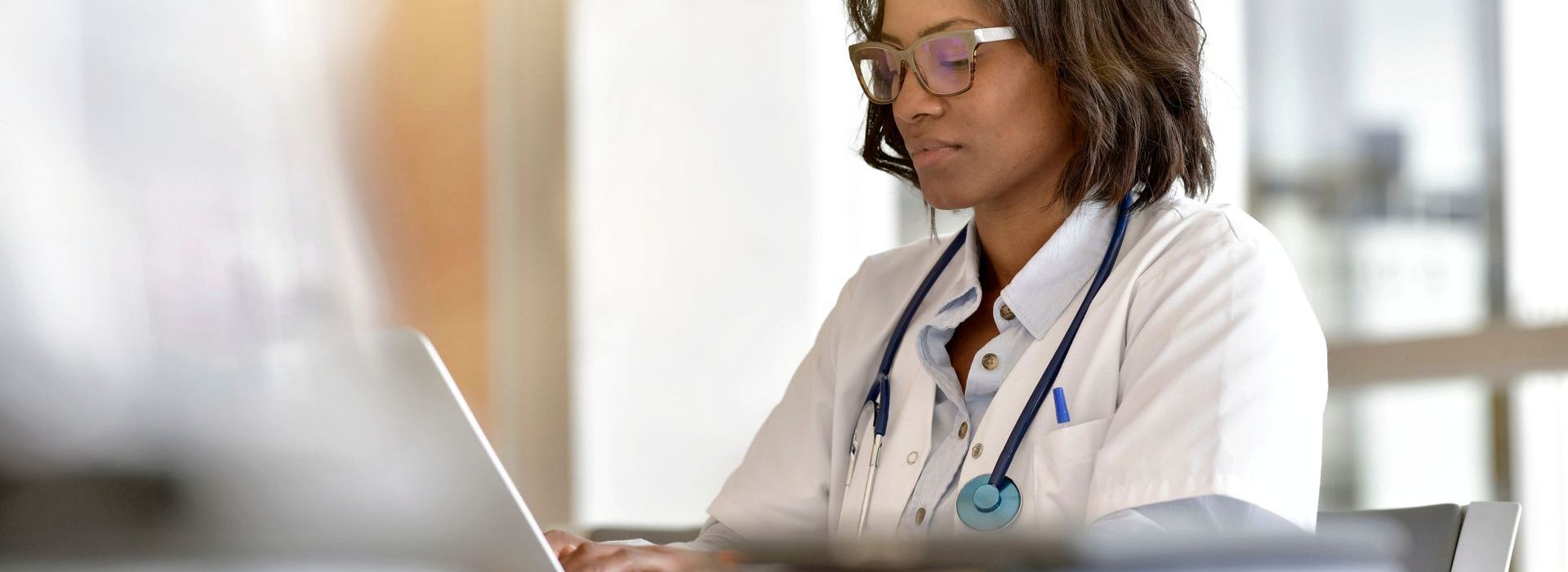 Une femme médecin noire avec un stéthoscope et des lunettes travaille sur un ordinateur portable dans un bureau.