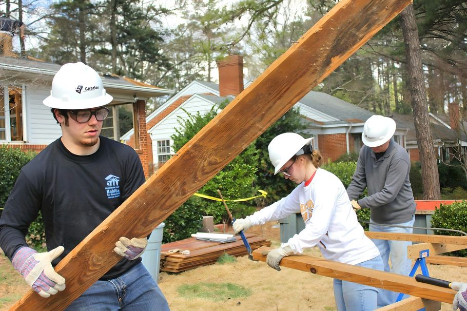 Three workers in white hard hats carry a long wooden beam at a construction site.