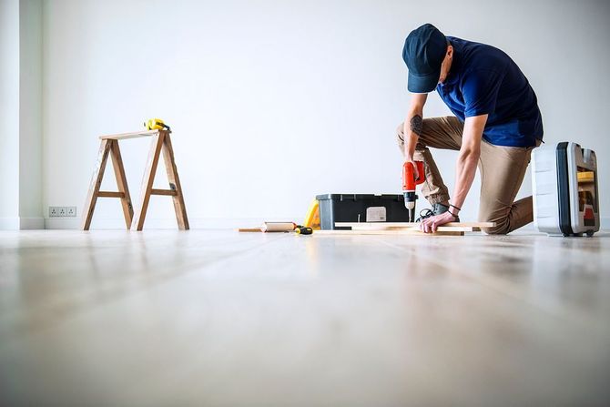 Person using a drill while kneeling on a floor in a bright room, with tools and a ladder nearby