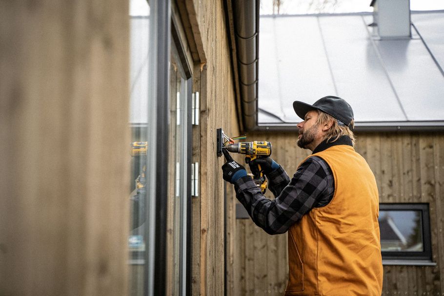 Worker in orange vest drilling into an exterior wall beside a window.